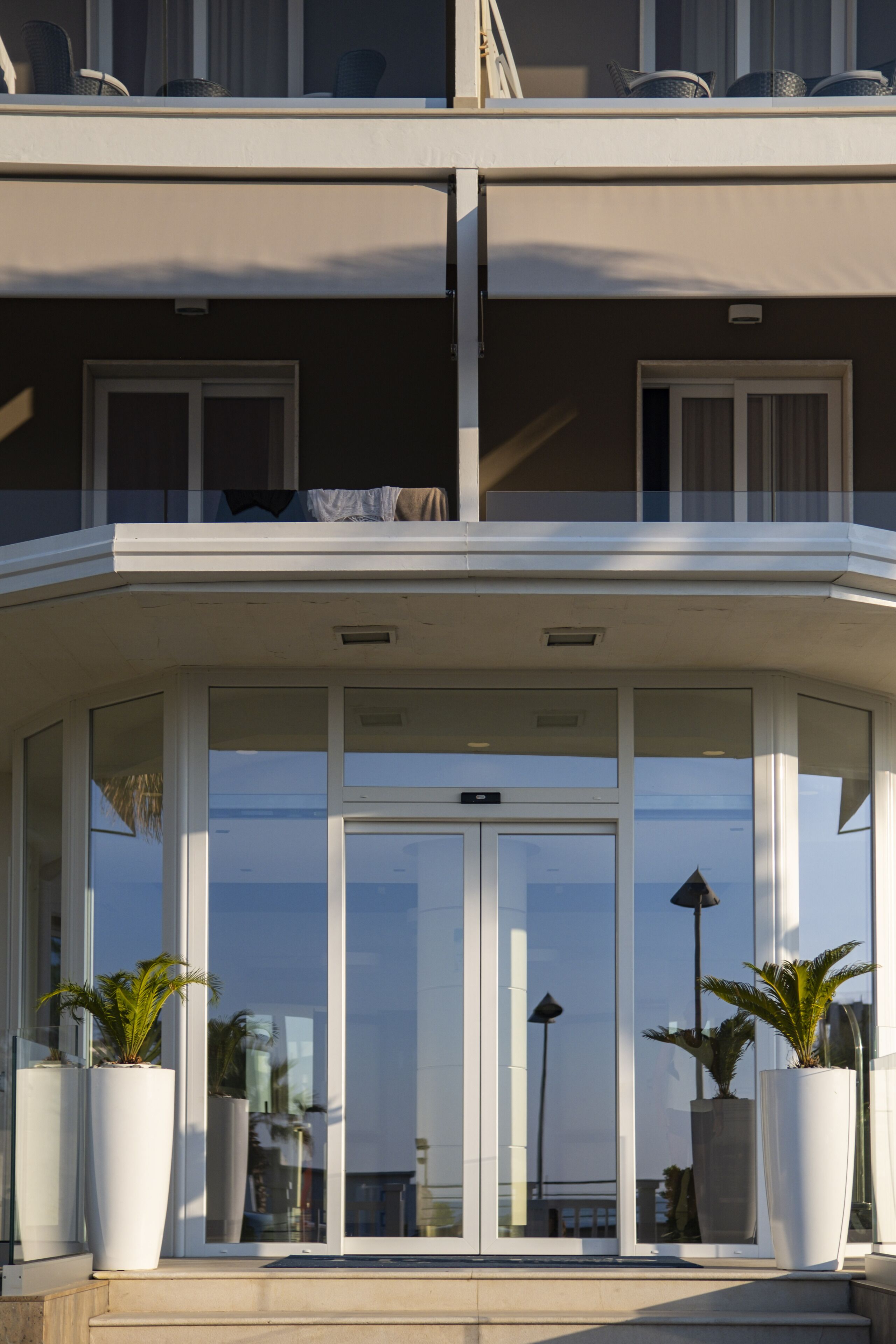 Un edificio moderno con ingresso in vetro, grandi piante in vaso e un balcone con sedie.