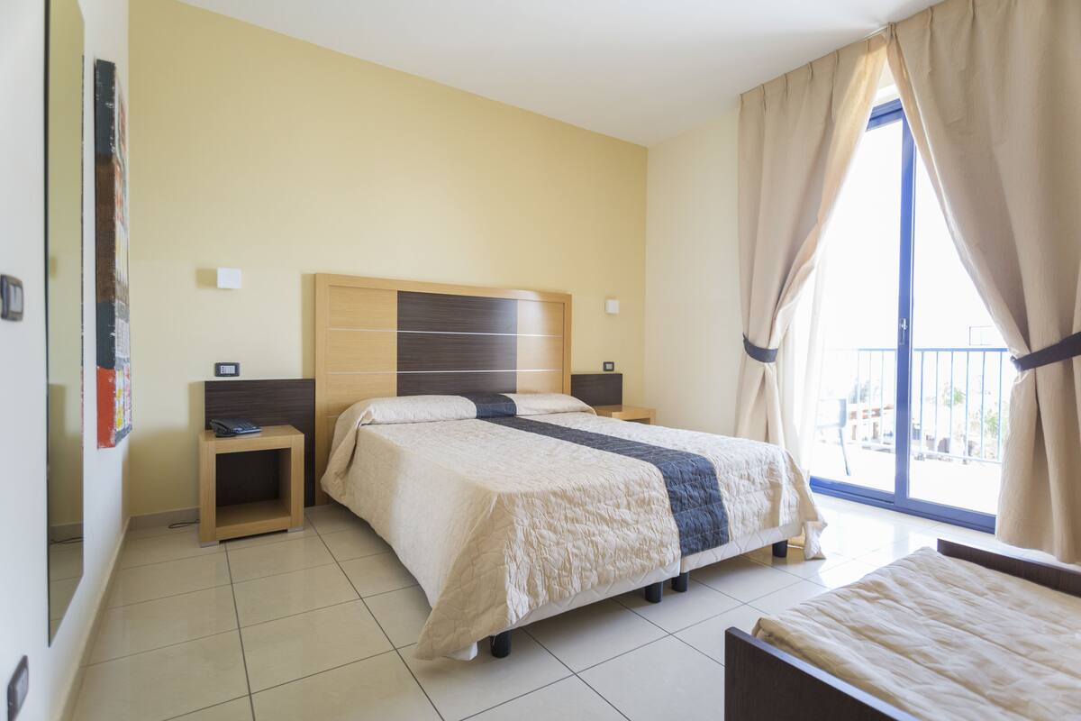 Une chambre d’hôtel avec un lit, des tables de chevet, une petite table de nuit en bois, une fenêtre avec des rideaux et une vue sur l’extérieur.