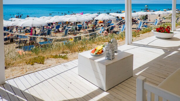 Une terrasse au bord de la plage, avec des meubles blancs, une lanterne blanche et un vase rempli de fleurs.