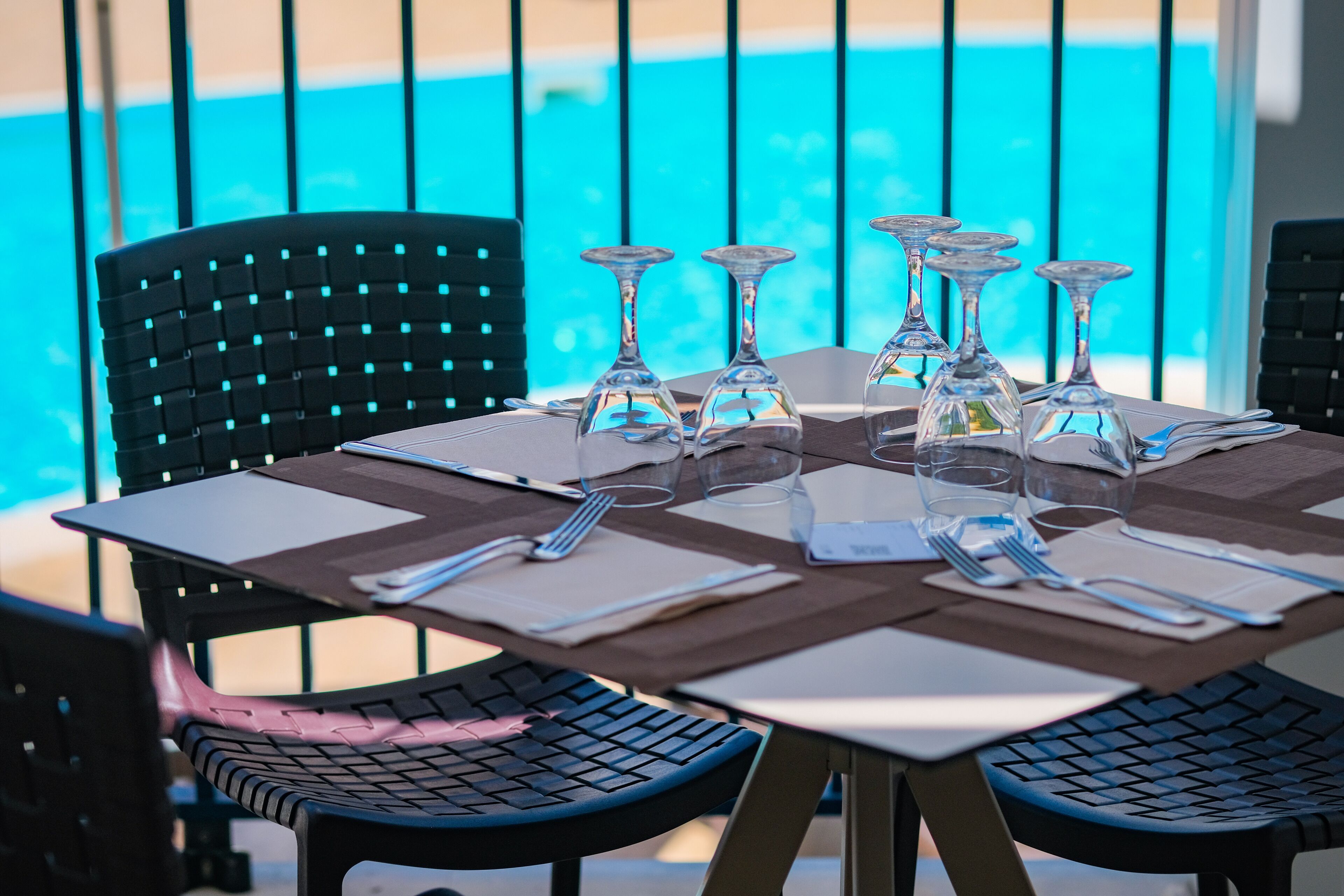 Mesa de comedor lista con varias copas de vino, cubiertos y servilletas, ubicada cerca de una zona de piscina con sillas.