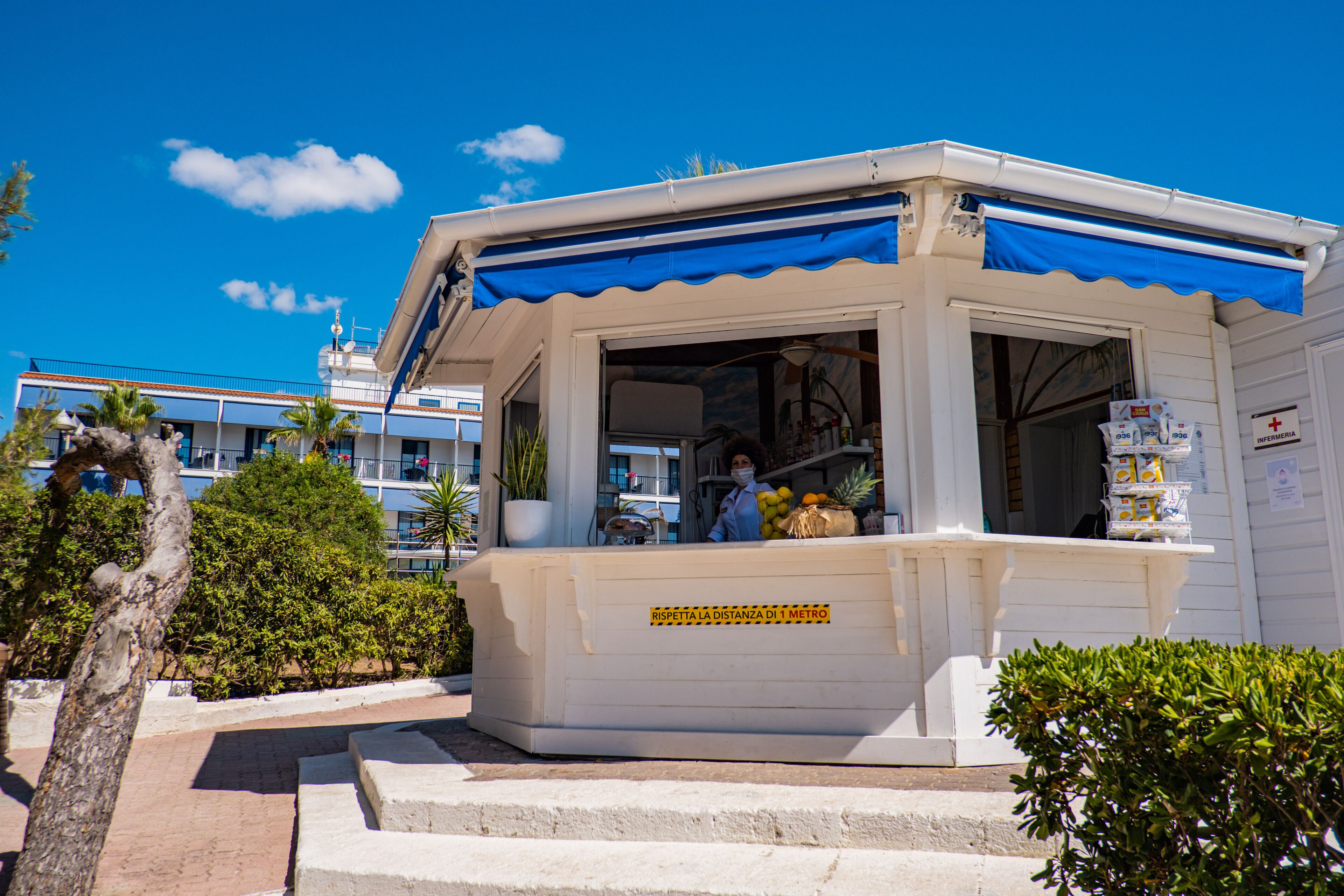 Un petit bâtiment blanc avec un auvent bleu, une affiche indiquant les mesures de distanciation sociale, et une personne à l’intérieur.