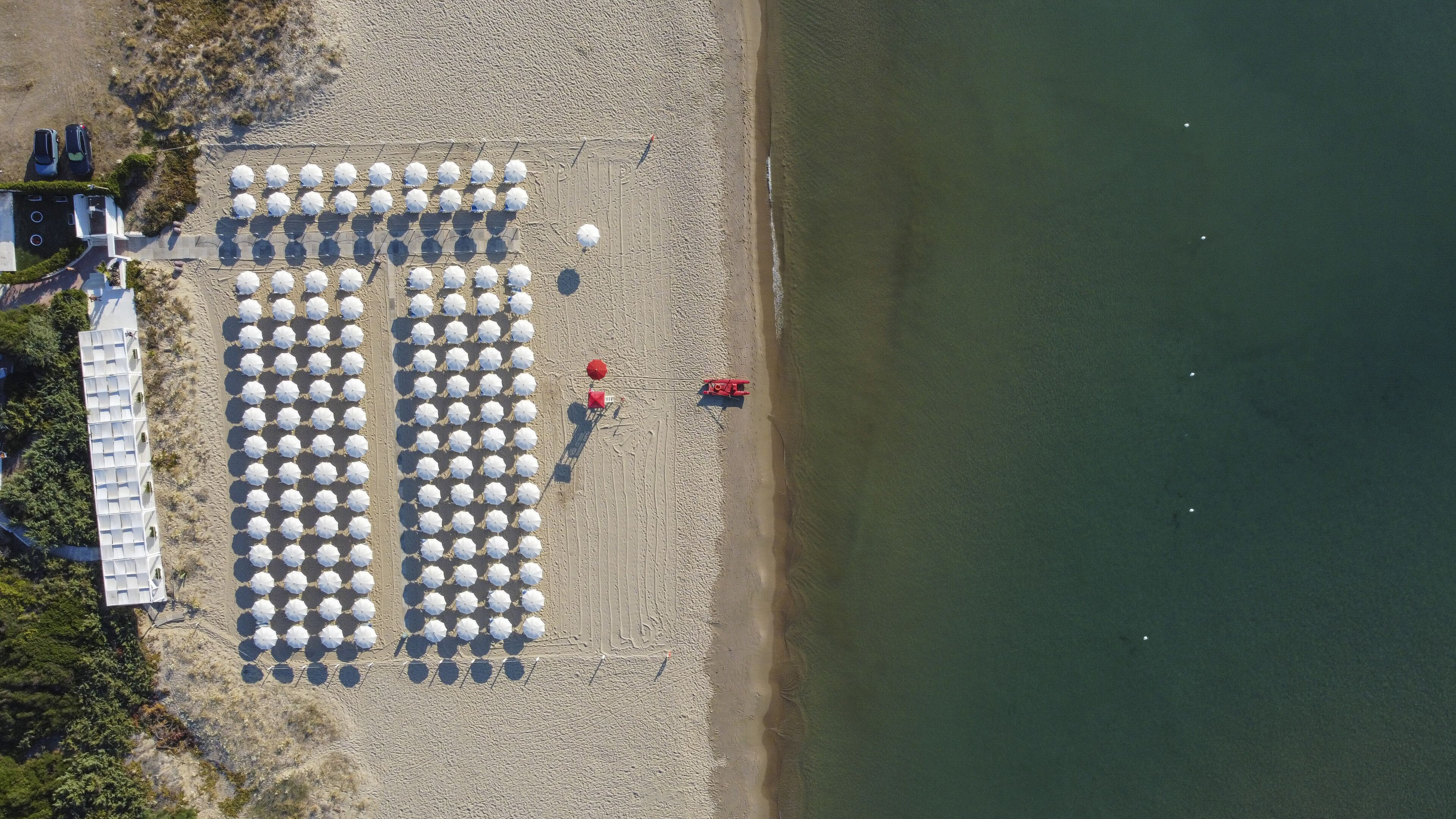 An aerial view of a beach with rows of white umbrellas, a red lifeguard tower, and a building with a white facade.