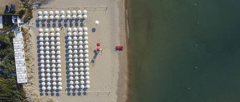 Vista aérea de una playa con hileras de sombrillas blancas, una torre roja de salvavidas y un edificio con fachada blanca.