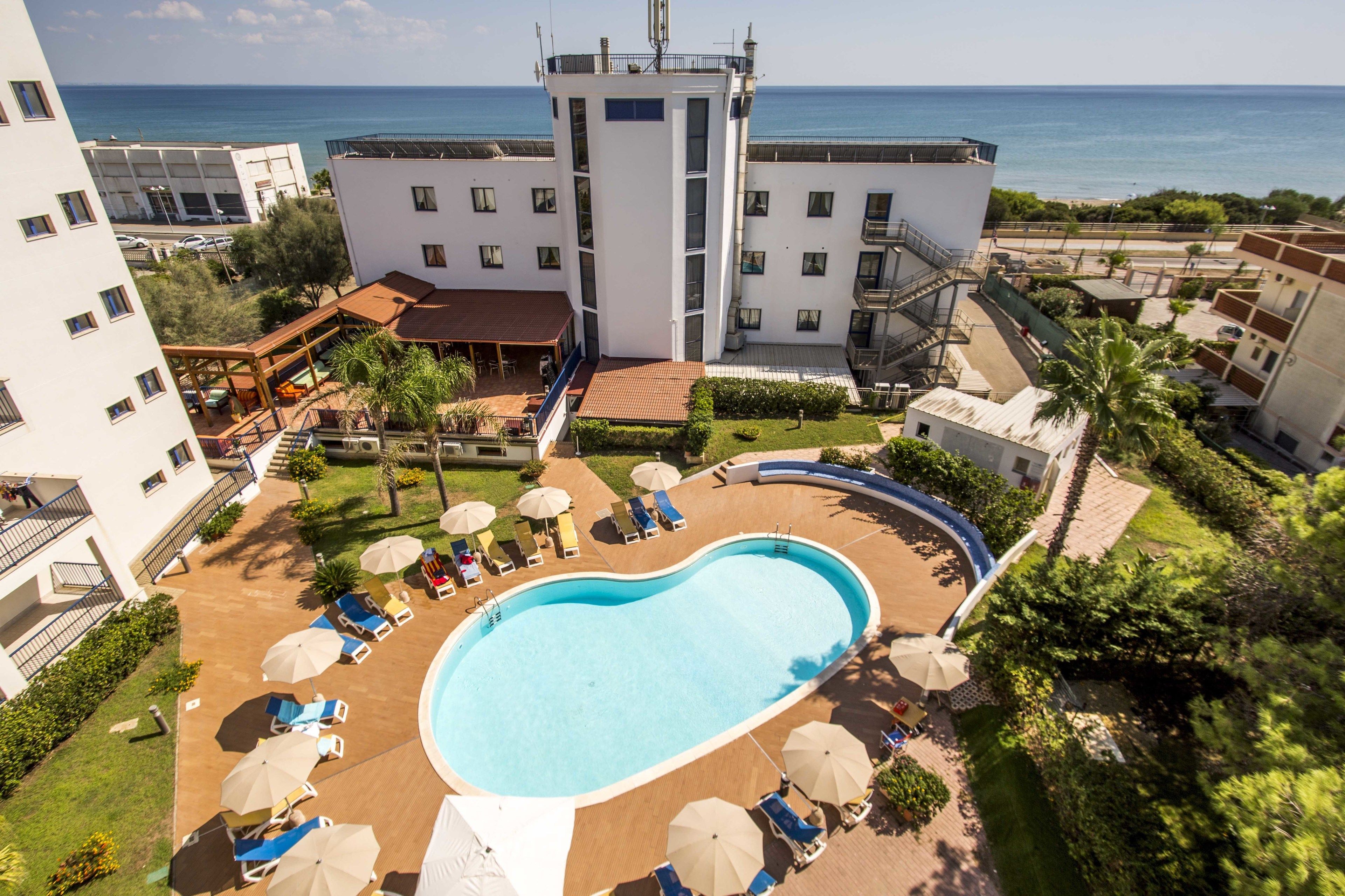 Hotel met een zwembad, omringd door ligbedden en parasols, met uitzicht op zee, en een gebouw met meerdere verdiepingen en balkons.