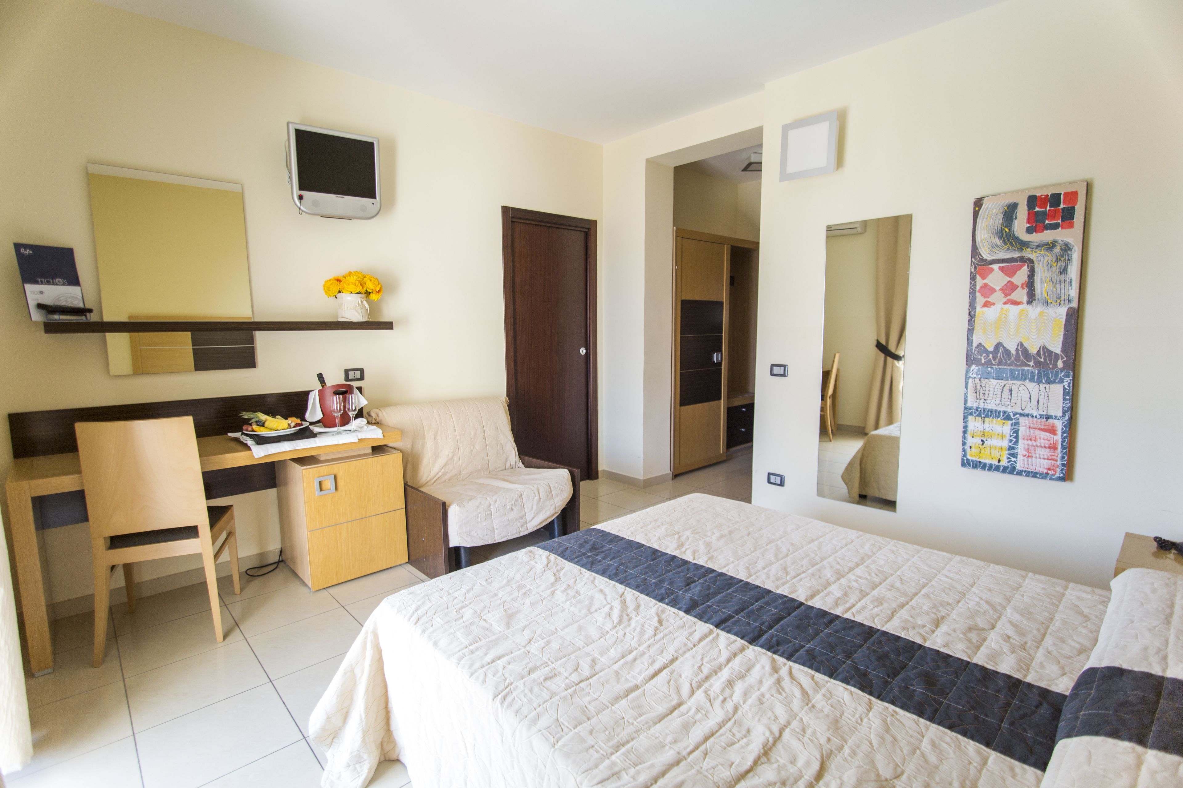 A hotel room with a bed, a desk with a chair, a small table with a vase of flowers, a television, and a door leading to another room.