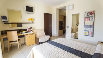 A hotel room with a bed, a desk with a chair, a small table with a vase of flowers, a television, and a door leading to another room.