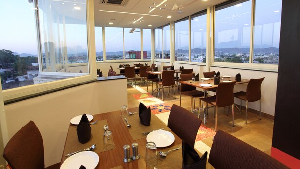 A dining area with tables set for four, large windows offering a city view, and a ceiling fan. - Hotel Dayal (Udaipur)