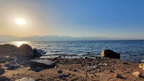 A rocky beach with large boulders and a clear view of the sea and distant mountains under a bright sun.