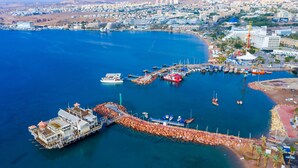 An aerial view of a coastal marina with boats docked, a large cruise ship, and a cityscape in the background.