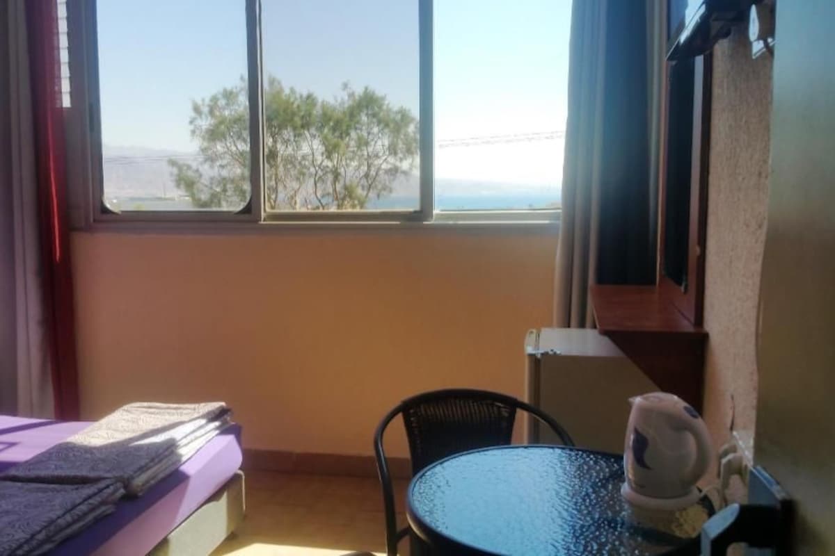 A room with a bed, a small table, a chair, and a window with a view of trees and the horizon.