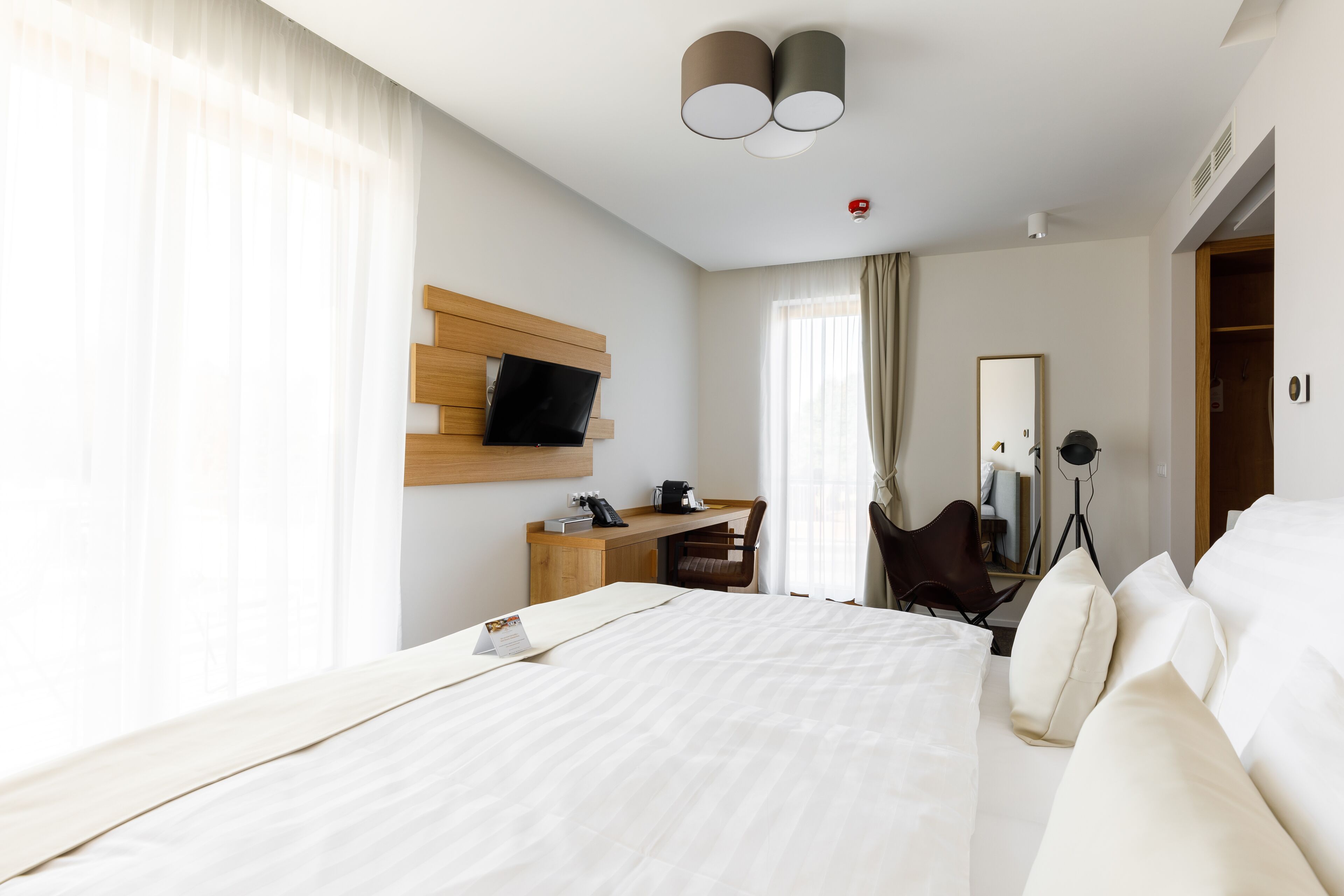 A modern hotel room with a large bed, a desk, a chair, a TV mounted on the wall, and a wardrobe.