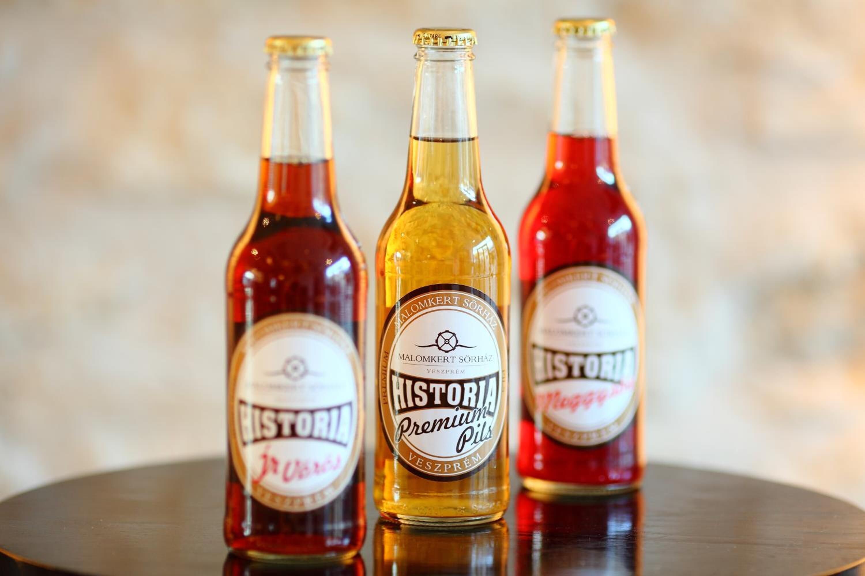 Three bottles of Historía Premium Pils on a reflective surface.