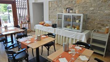 A dining area with tables set for breakfast, a buffet with food and drinks, and a stone wall backdrop.