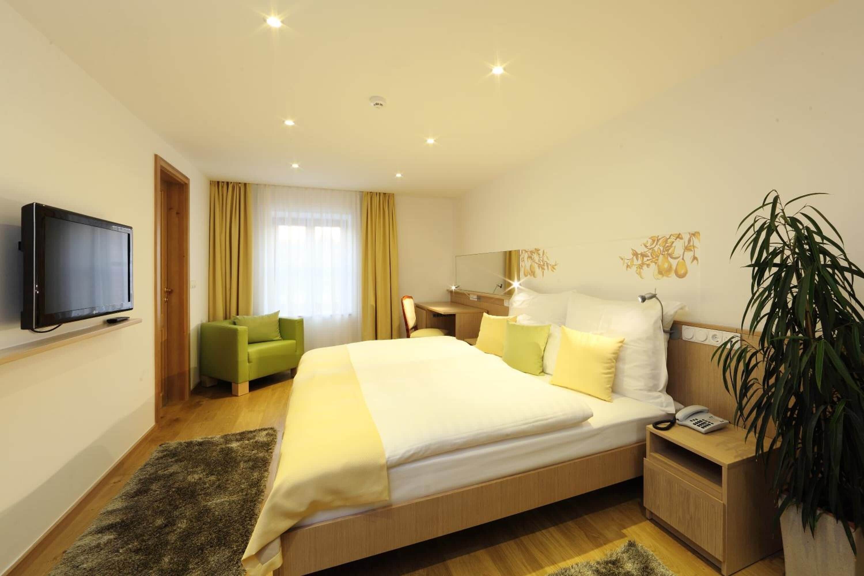 A modern hotel room with a large bed, a green armchair, a desk with a lamp, and a television on the wall.