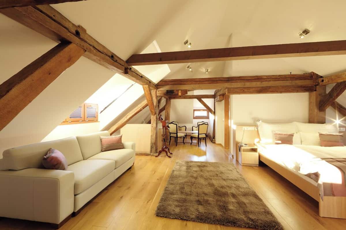 A loft bedroom with a sloped ceiling, wooden beams, a sofa, a bed, and a dining table.
