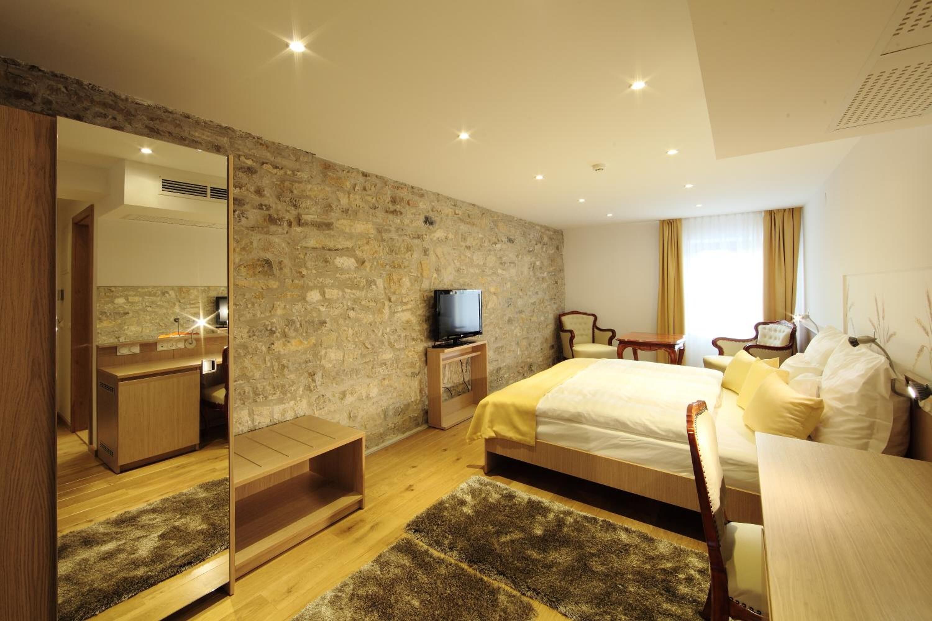 A modern hotel room with a stone accent wall, a wooden bed, a television, and a desk with a chair.