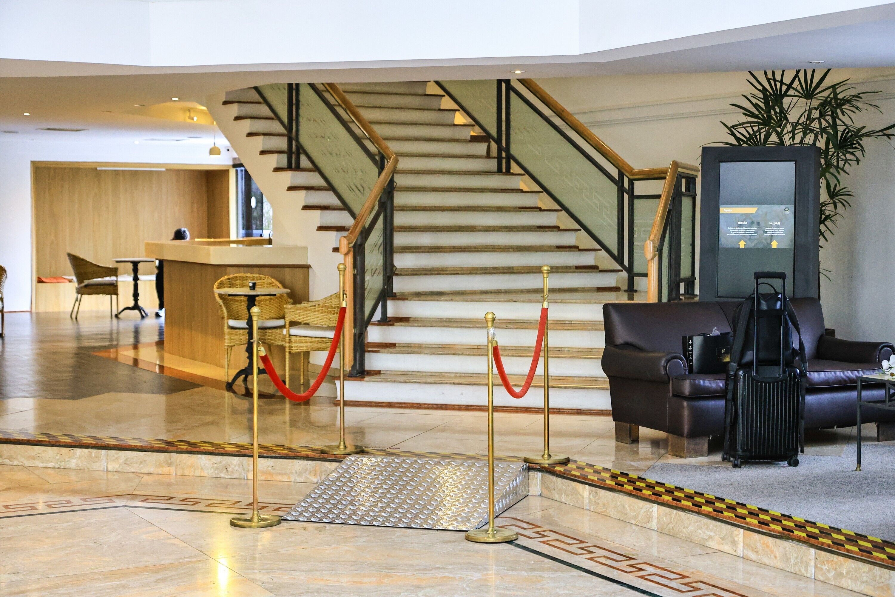 A hotel lobby with a reception desk, a staircase, a seating area, and a plant.