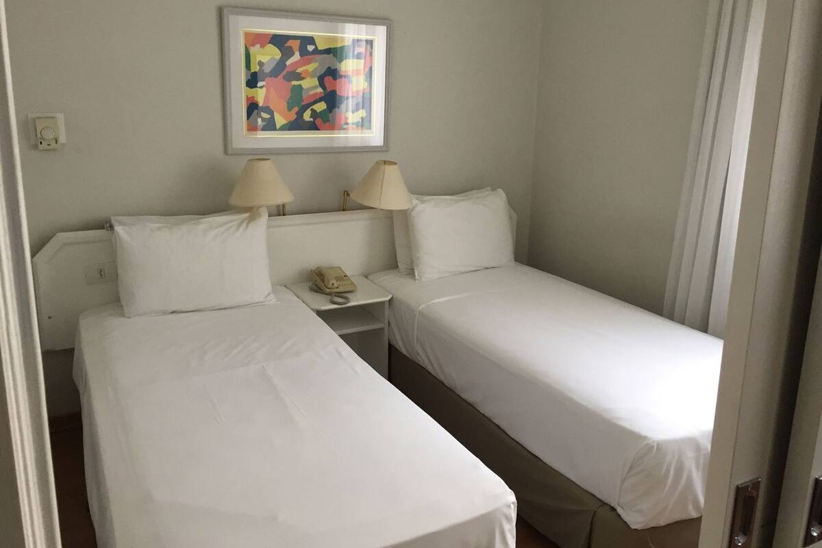 A hotel room with two single beds, a bedside table with a lamp, a phone, and a framed abstract painting on the wall.