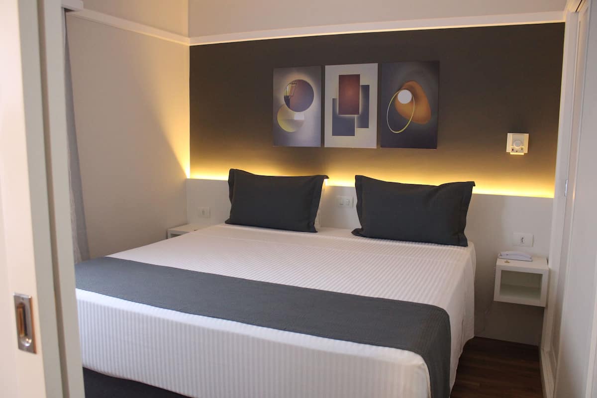 A neatly made bed with a white and gray bedspread, two dark pillows, and a bedside table with a lamp.