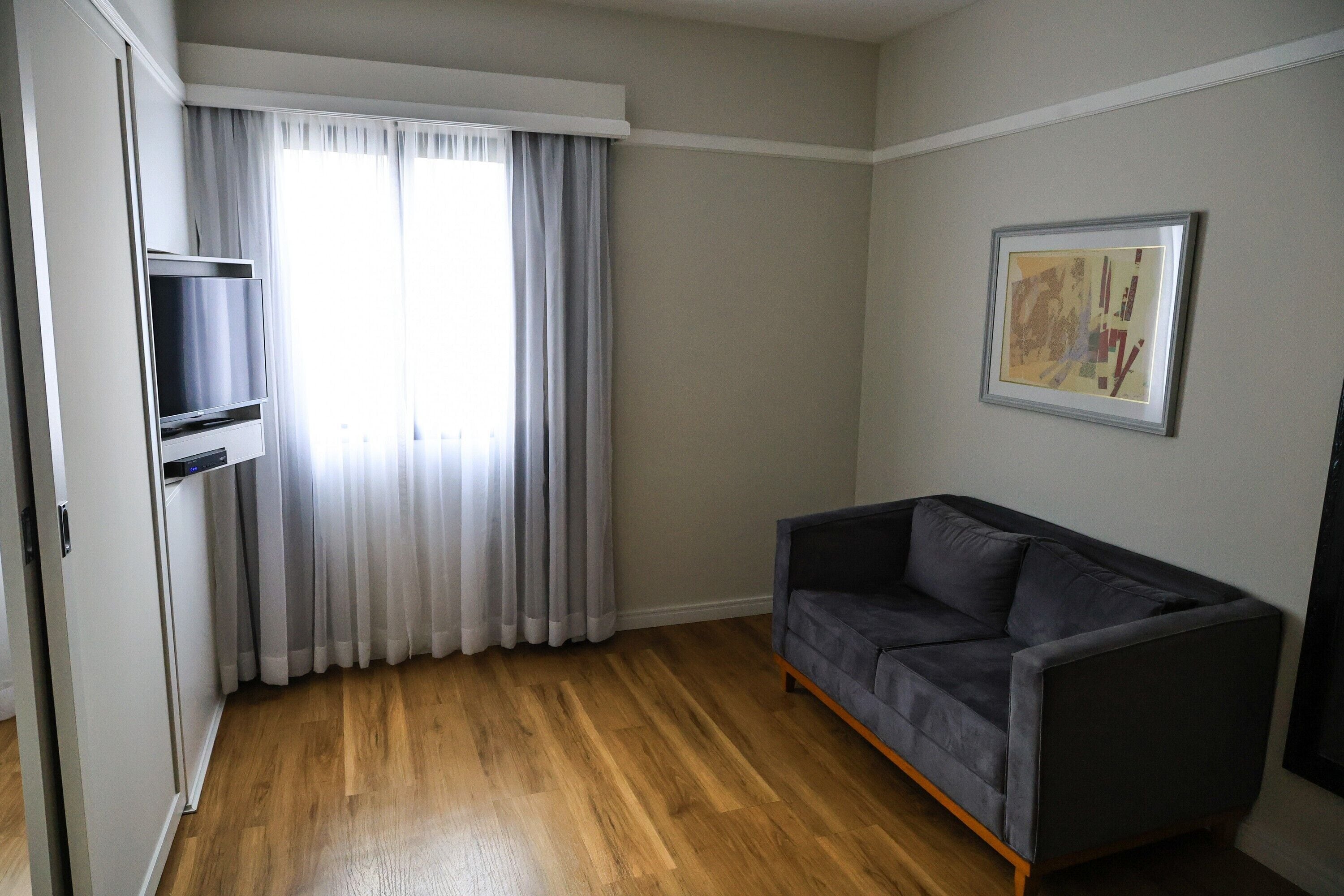 Una habitación con sofá gris, suelo de madera, ventana con cortinas e imagen enmarcadas en la pared.