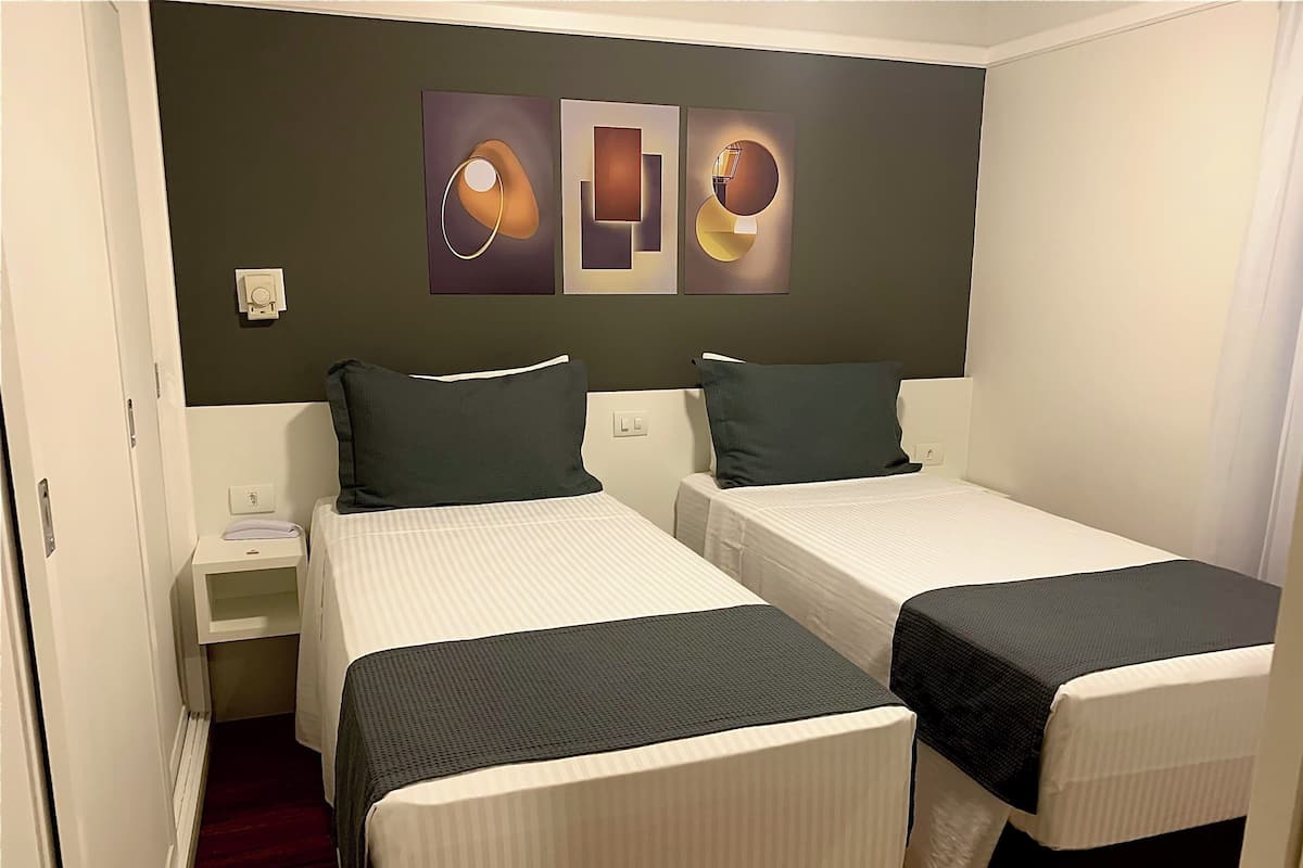 A hotel room with two single beds, a small bedside table with a lamp, and a wall-mounted artwork above the beds.