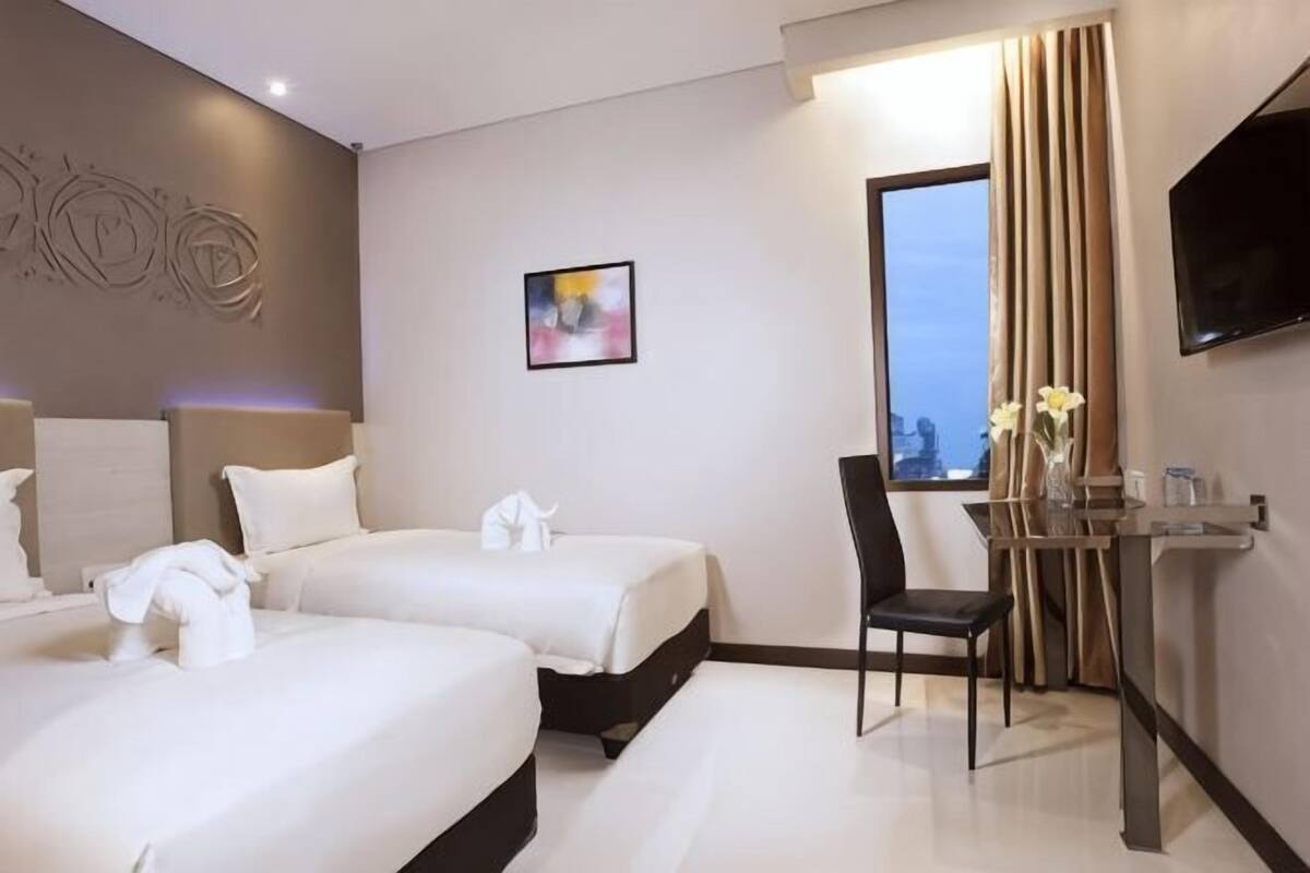 A hotel room with two beds, a desk, a chair, a TV, and a window with curtains.