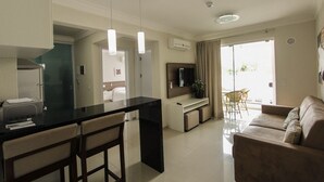 A modern kitchen with a black countertop, two bar stools, a dining table, and a TV mounted on the wall. There is a sofa and a chair in the living area, and a balcony with a view. - Bombinhas Summer Beach Hotel & Spa (Bombinhas)