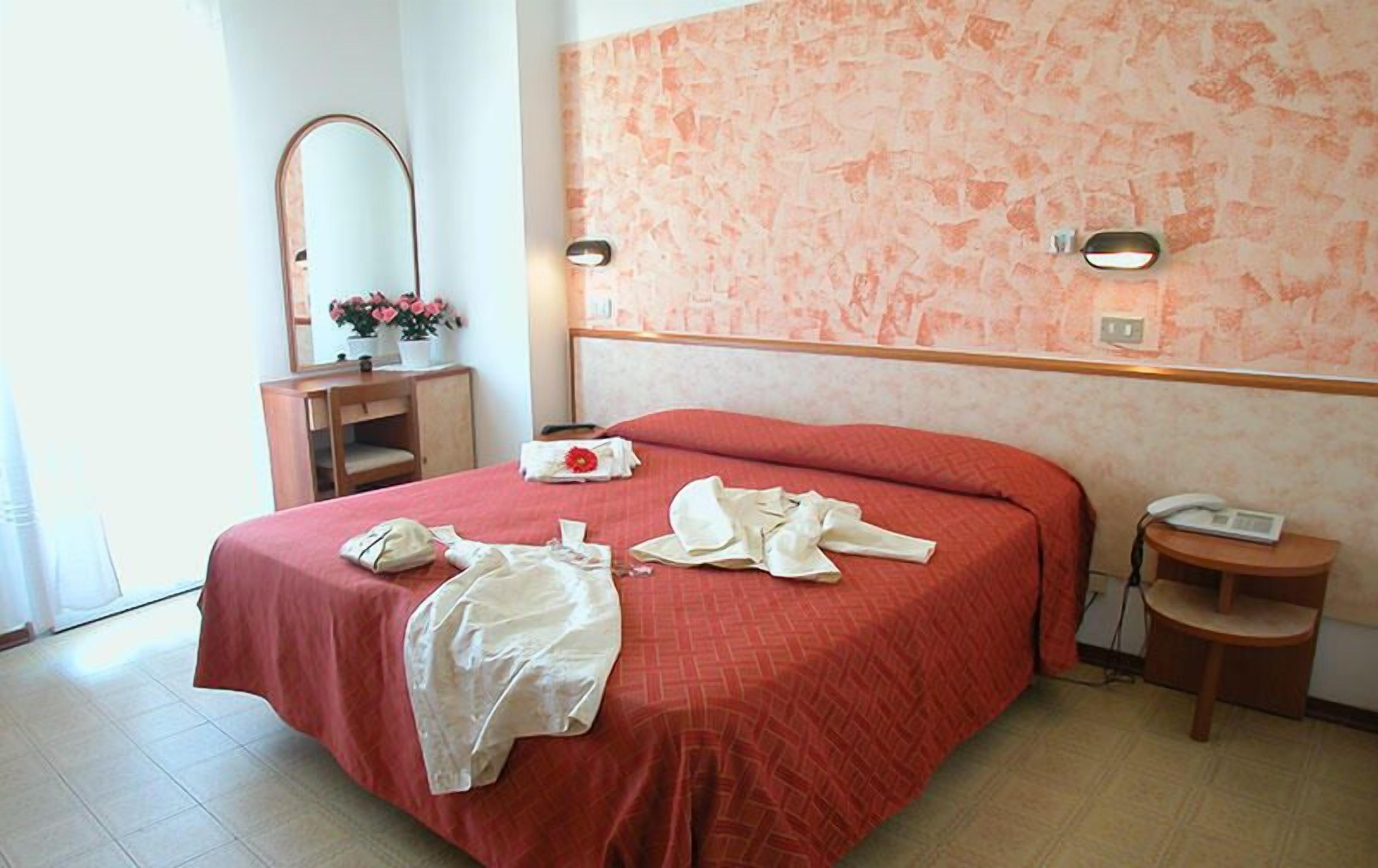 A hotel room with a bed, a bedside table, a mirror, and a wall-mounted light fixture.