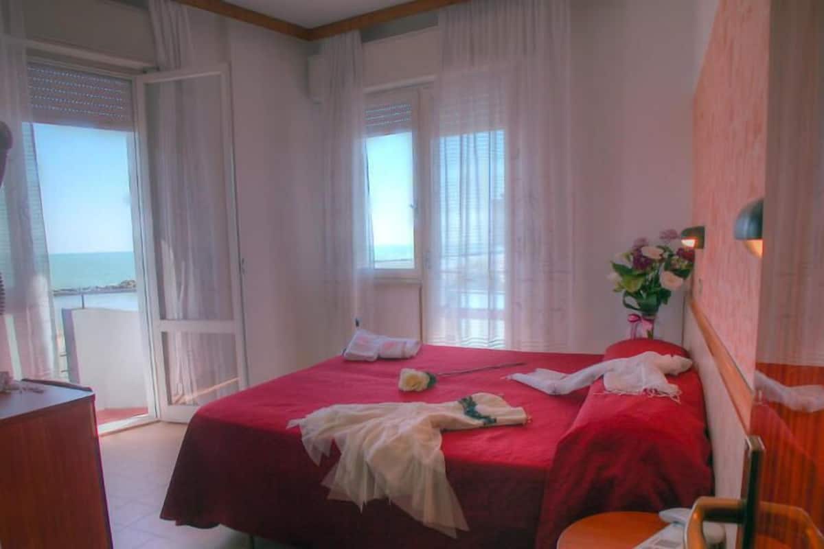 Ein Schlafzimmer mit rotem Bettbezug, einem Holz-Kopfschild, einem Nachttisch und Blick auf das Meer durch das Fenster.