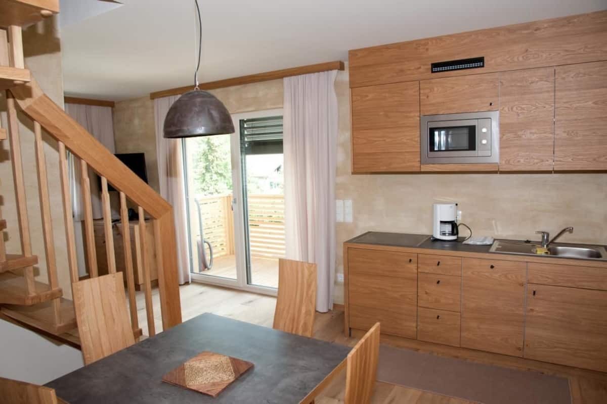 A modern kitchen with wooden cabinets, a microwave, and a coffee maker. There is a dining area with a table and chairs, and a staircase leading to another level.