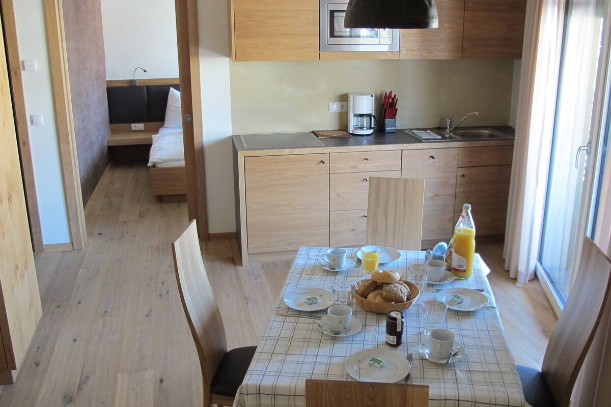 A compact kitchen with wooden cabinets, a dining table set for breakfast, and a bed in the adjacent room.