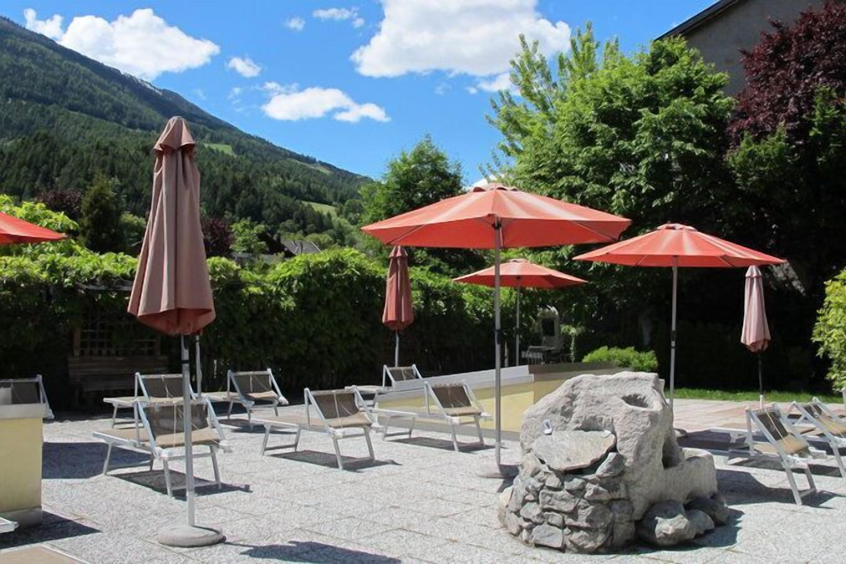 Un espace extérieur aménagé avec des fauteuils confortables et des parasols rouges, le tout se fondant dans un décor de verdure, avec des arbres et une montagne en toile de fond.