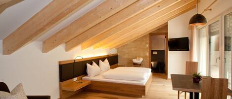 A modern bedroom with a sloped ceiling, a bed with white linens, a bedside table, a wooden nightstand, a small seating area with a couch, and a dining table with chairs.