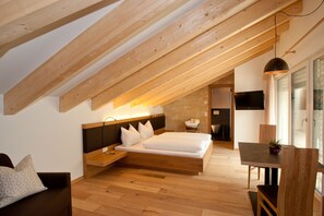 Ein modernes Schlafzimmer mit schrägem Dach, einem Bett mit weißer Bettwäsche, einem Nachttisch, einem Holzbeistelltisch, einer kleinen Sitzecke mit einer Couch und einem Esstisch mit Stühlen.