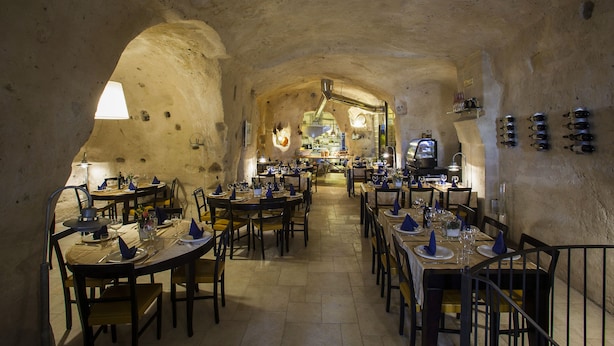 A restaurant with tables set for dining, a kitchen in the background, and a rustic stone wall.