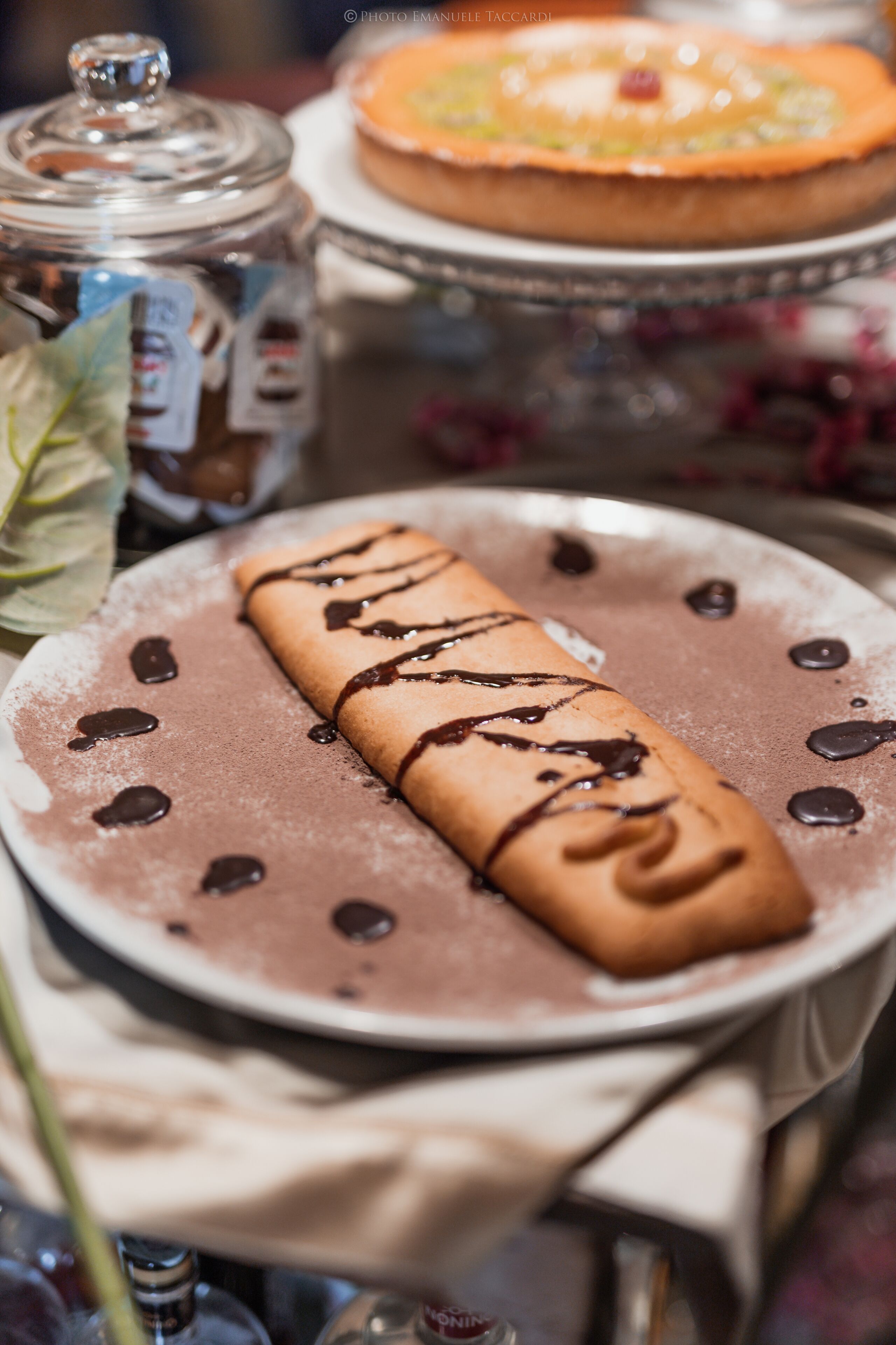 A dessert plate with a chocolate-dipped crepe, a jar of jam, and a cake on a table.