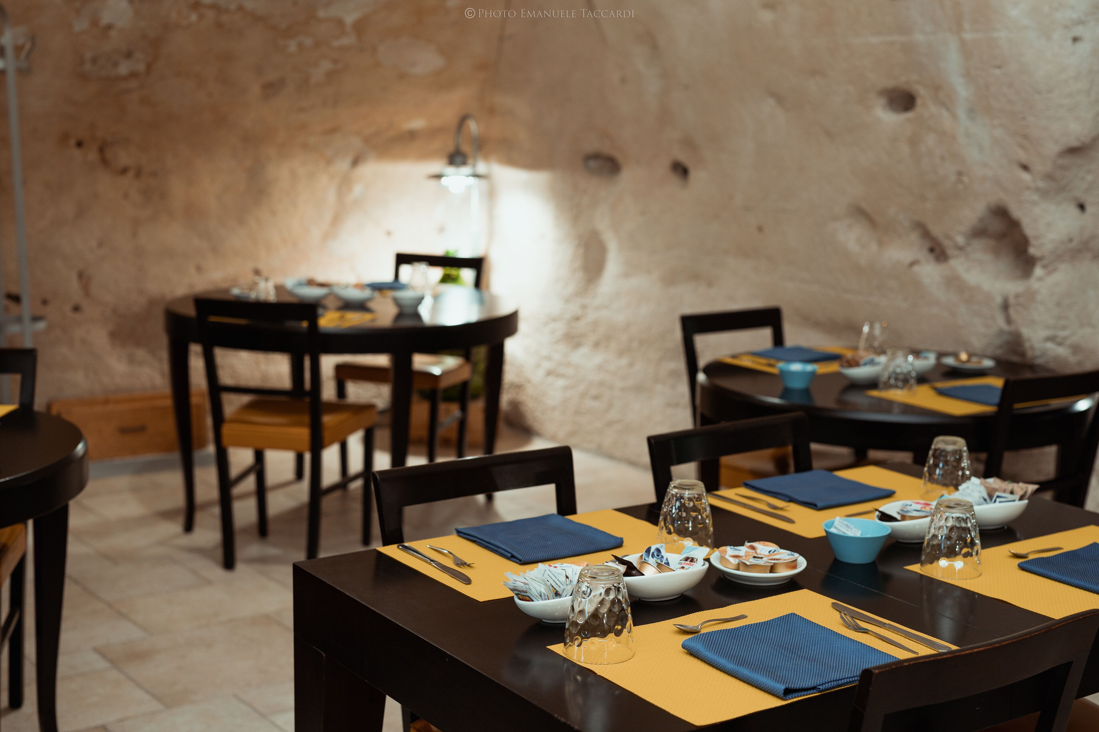 A dining area with tables set for two, featuring a rustic stone wall in the background.