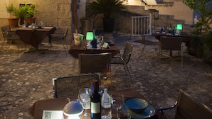 An outdoor dining area with stone tables and chairs, potted plants, and a view of a historic building with a bell tower in the background.