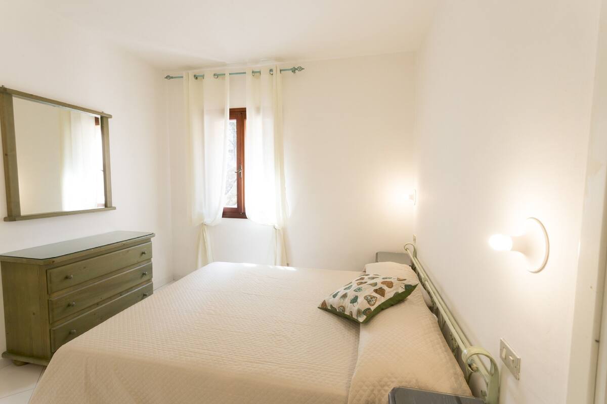 Um quarto com cama, penteadeira, janela com cortinas e luminária na parede.