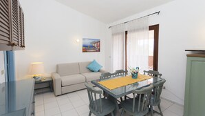 Un comedor con una mesa de vidrio, sillas de madera, un sofá con un cojín azul, un cuadro en la pared y una ventana con cortinas translúcidas.