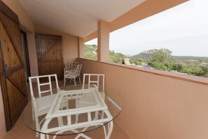 Een balkon met een glazen tafel en stoelen, een houten deur en uitzicht op groen en heuvels.