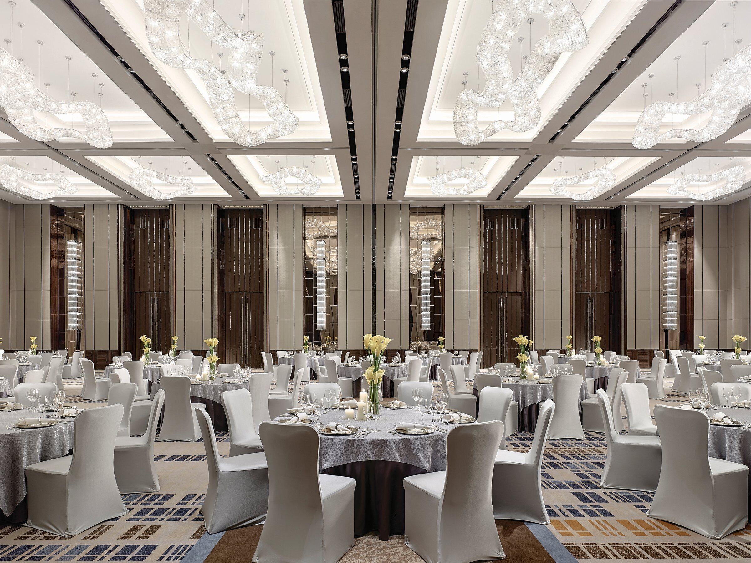 A spacious banquet hall with round tables, white chairs, and chandeliers.