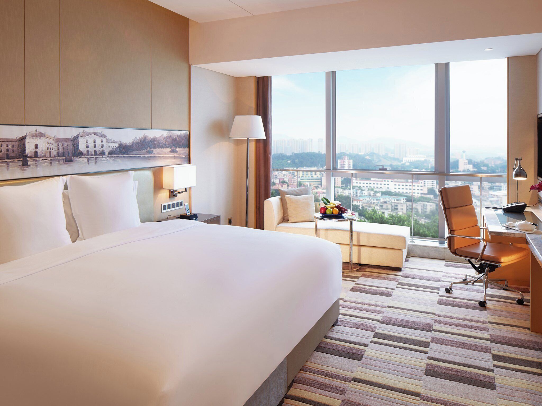 A hotel room with a large bed, a desk with a chair, a small table with fruit, and a view of a cityscape through large windows.
