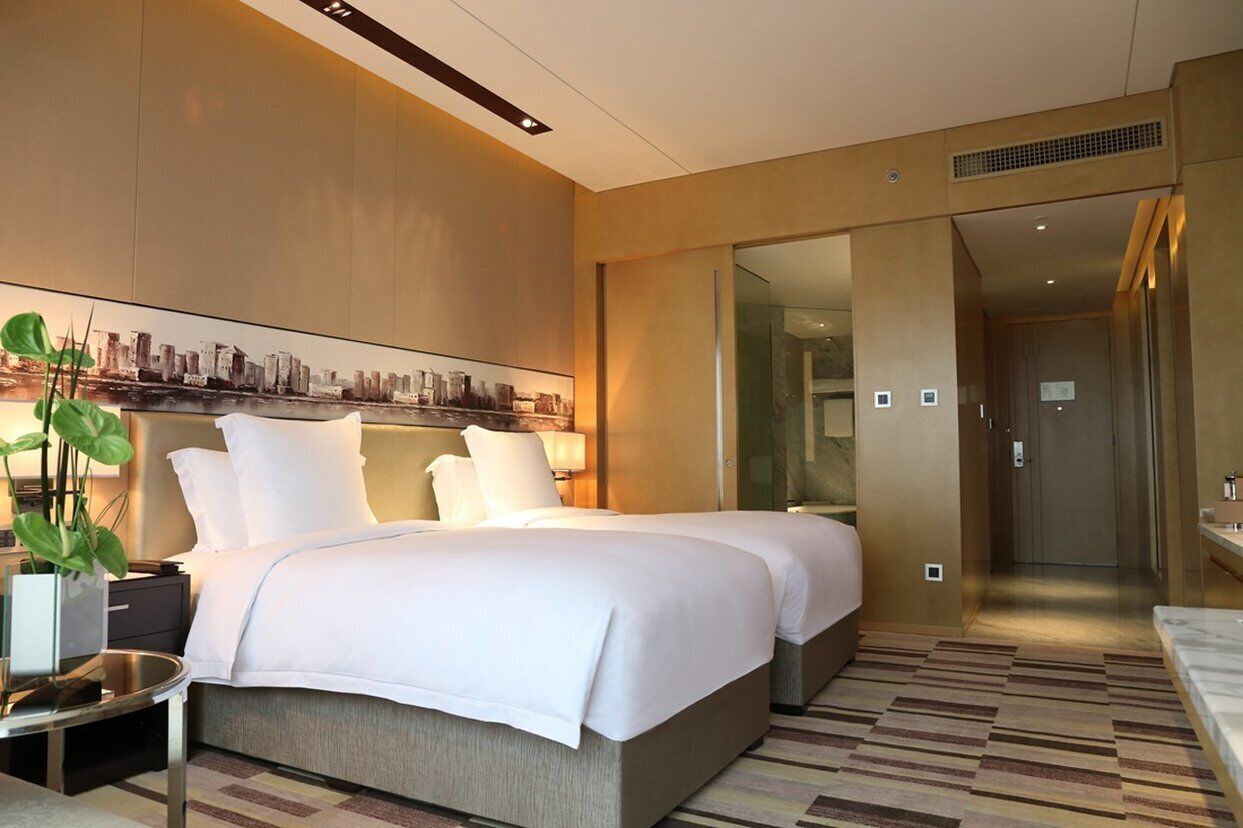 A modern hotel room with a large bed, a bedside table with a lamp, a potted plant, and a cityscape artwork on the wall.