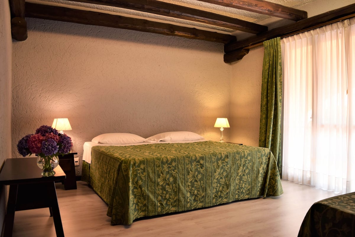 Ein Zimmer mit einem Bett, zwei Nachttischlampen, einem kleinen Tisch mit Blumenvase und einem Fenster mit Vorhängen.