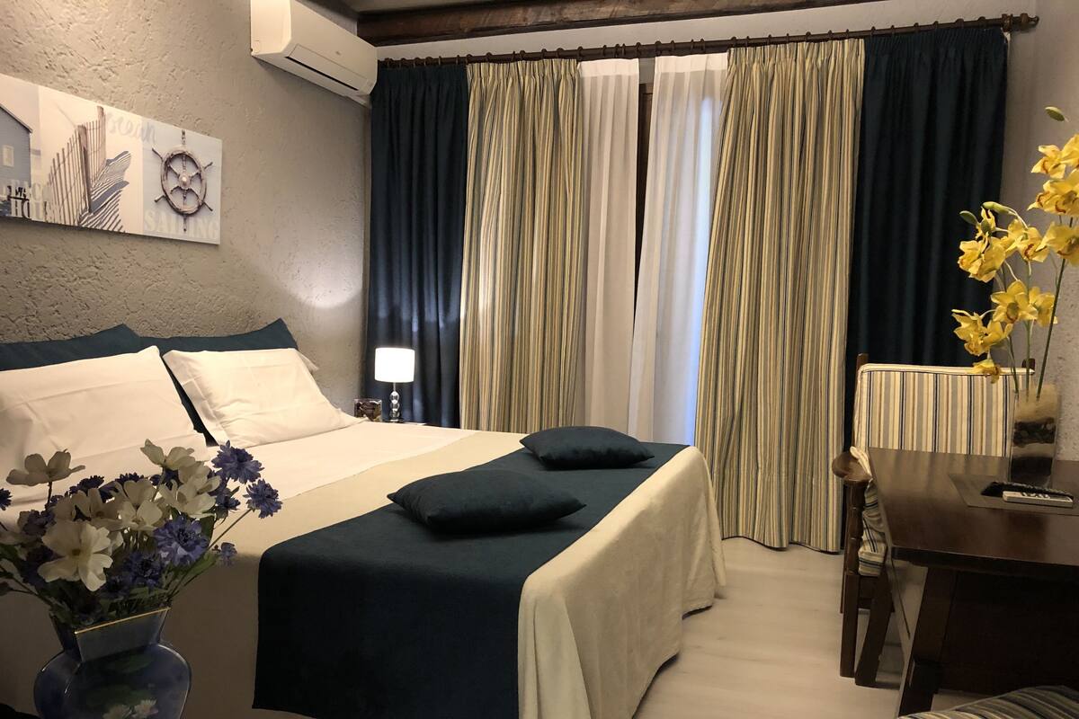 Ein Hotelzimmer mit Bett, Nachttisch, Lampe, Blumenvase und Blick ins Freie durch Vorhänge.
