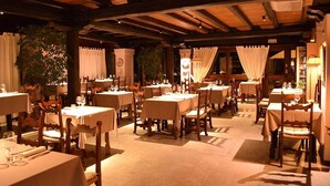 En restaurant med flere borde og stole, træbjælker i loftet og gardiner ved vinduerne.