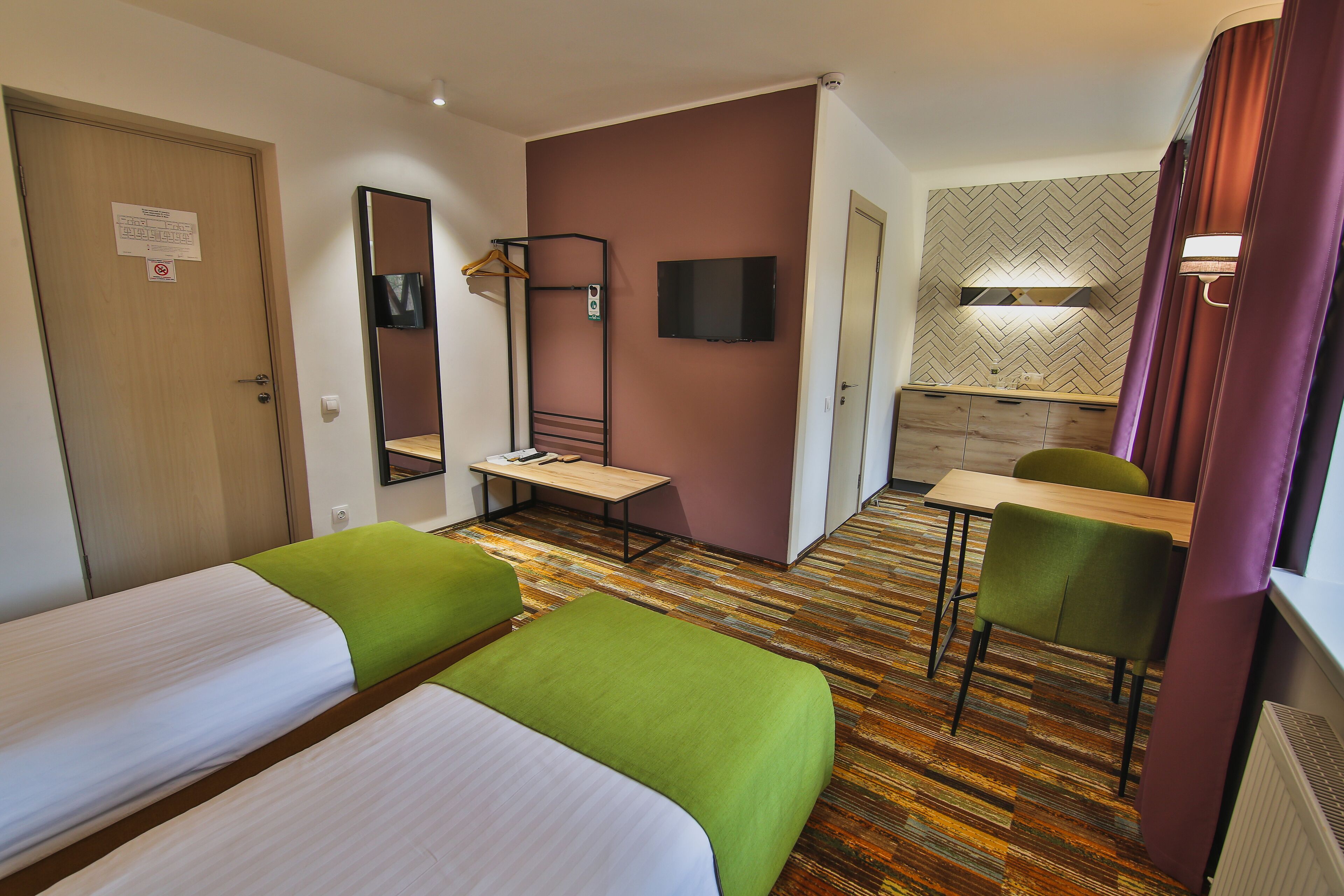 A hotel room with two single beds, a desk, a chair, a TV, and a herringbone-patterned wall.