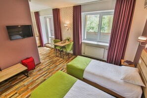 Ein Hotelzimmer mit zwei Betten, einem Schreibtisch, einem Stuhl, einem Fernseher und einem Fenster mit Vorhängen.