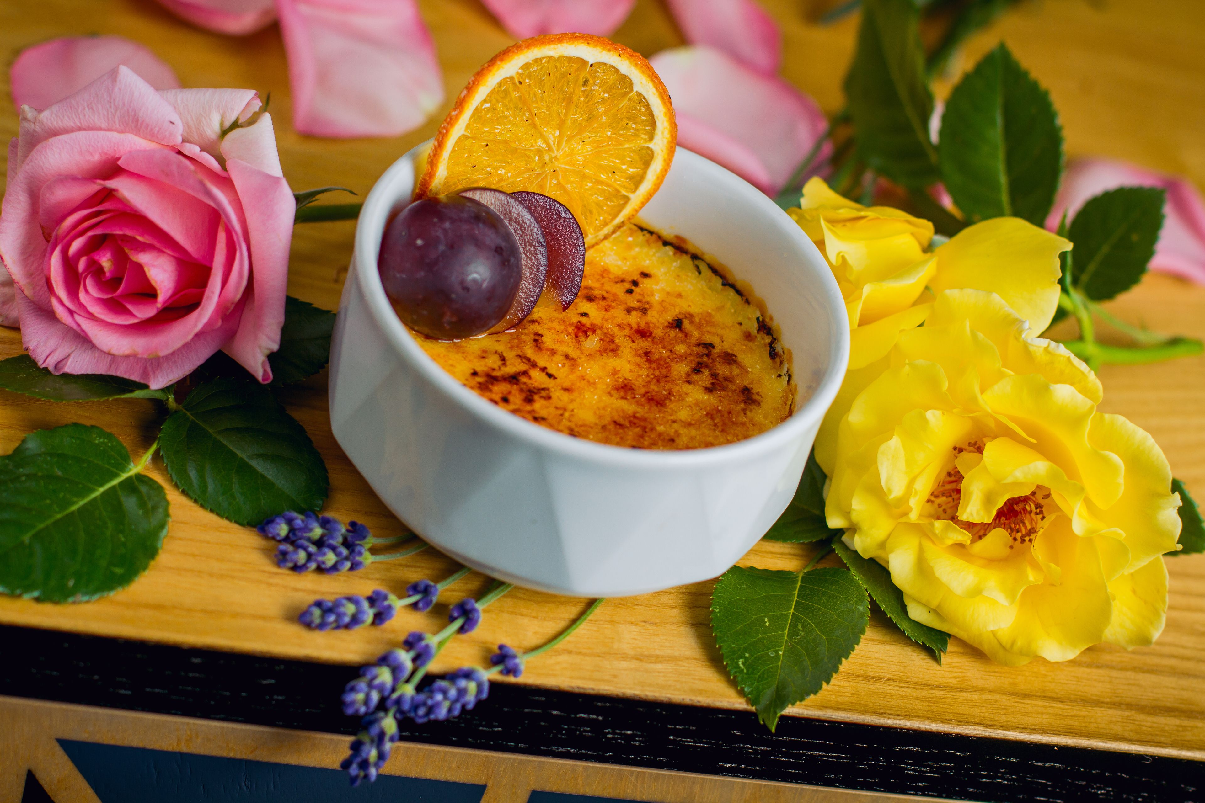 Un dessert in una ciotola bianca con una fetta d'arancia e un prugna sopra, circondato da rose rosa e gialle su un tavolo di legno.